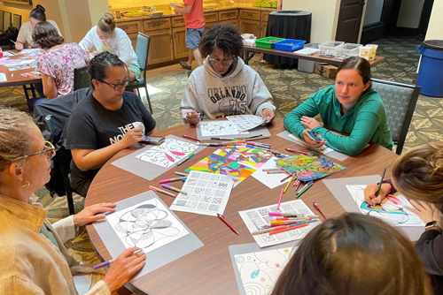 A group of women sit at a round table using markers to color on coloring sheets
