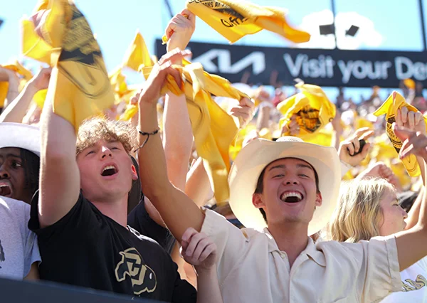 A group of CU students wearing black and gold and cheering at a large outdoor gathering