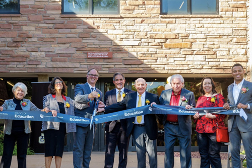 Several people stand smiling in front of the school of education building, they hold a long blue ribbon for the ribbon cutting ceremony