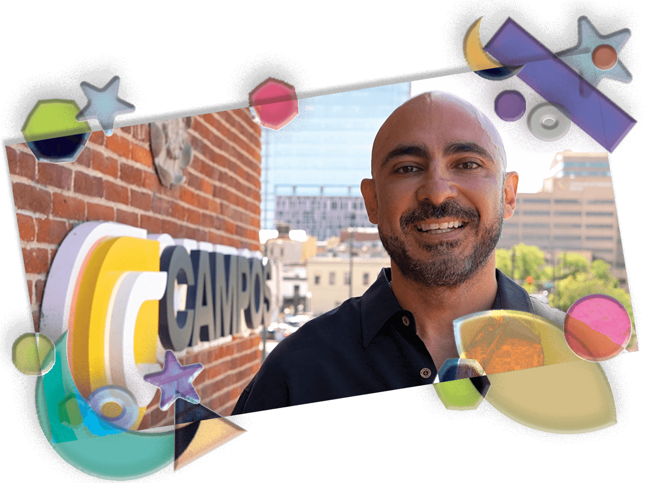 Marco Campos stands smiling jovially in front of a red brick wall with his company logo that reads "Campos." CU Denver is seen in the background.